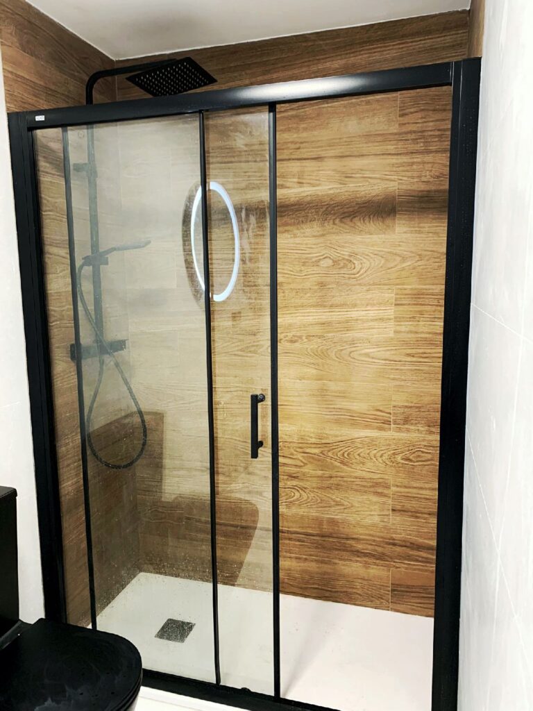 Zona ducha con mampara en negro y alicatado en simulación madera en el ámbito de baño